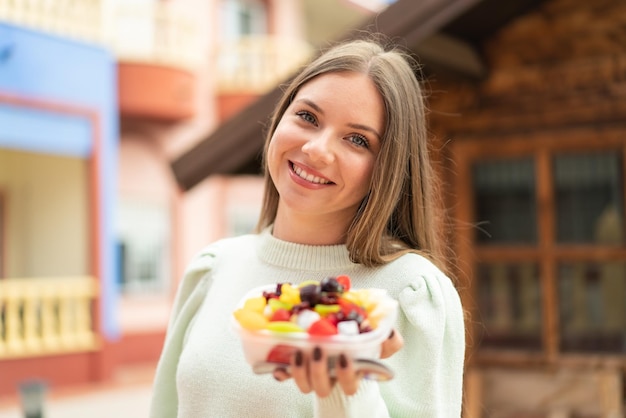 Jovem mulher bonita loira segurando uma tigela de frutas ao ar livre com expressão feliz