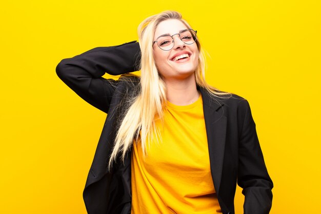 Foto jovem mulher bonita loira sorrindo alegremente e casualmente, levando a mão a cabeça com um olhar positivo, feliz e confiante contra parede amarela