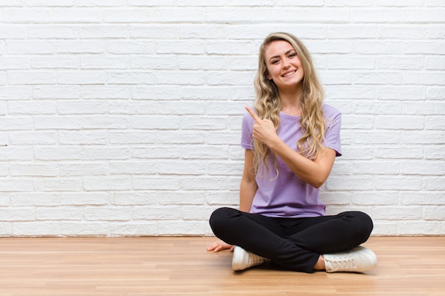 Jovem mulher bonita loira sorrindo alegremente, sentindo-se feliz e apontando para o lado e para cima, mostrando o objeto no espaço da cópia