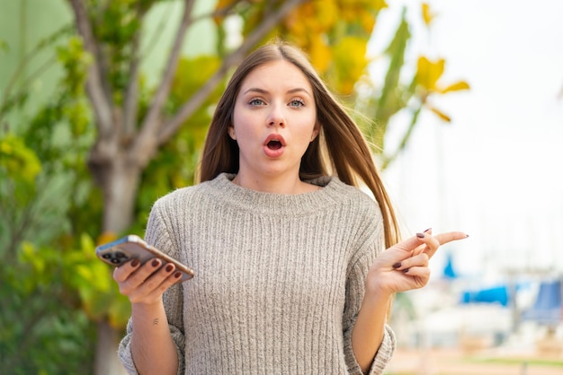 Jovem mulher bonita loira usando telefone celular ao ar livre surpreso e apontando para o lado