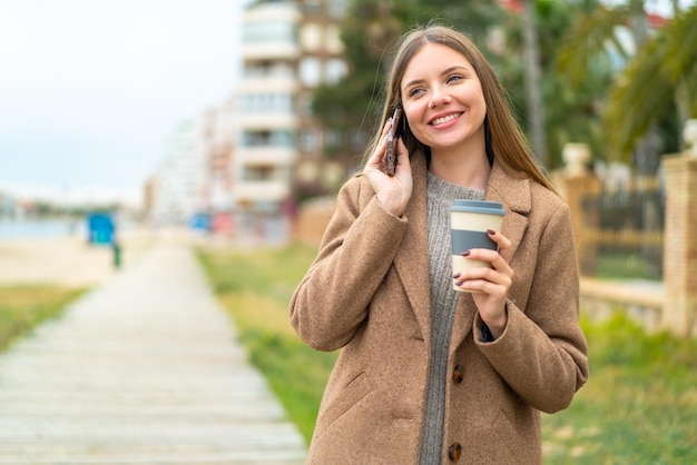 Jovem mulher bonita loira usando telefone celular e segurando um café com expressão feliz