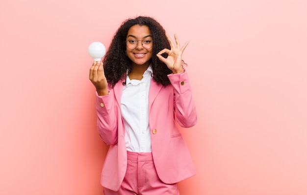 Jovem mulher bonita negra com uma lâmpada, tendo uma ideia contra o fundo da parede-de-rosa