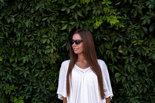 Jovem mulher bonita no vestido branco e óculos escuros sobre fundo verde arbusto. o conceito de alegria, facilidade e liberdade durante as férias. a garota está gostando do resto. foto de rua da moda.