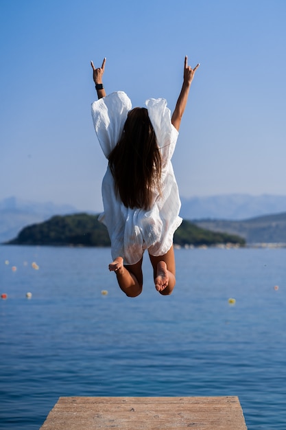 Foto jovem mulher bonita no vestido branco pulando no cais com fundo de vista mar. o conceito de alegria, facilidade e liberdade durante as férias. a garota está gostando do resto. conceito de liberdade