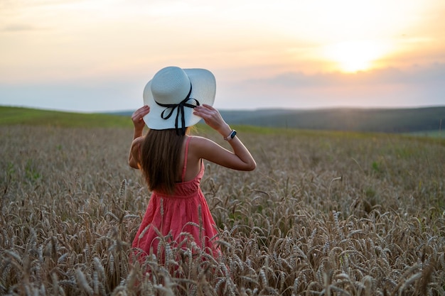 Jovem mulher bonita no vestido de verão vermelho e chapéu de palha em pé no campo de fazenda amarelo com trigo dourado maduro, aproveitando a noite quente.