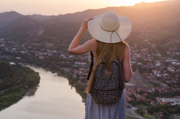 Jovem mulher bonita no vestido e chapéu, olhando o rio e o pôr do sol da cidade.