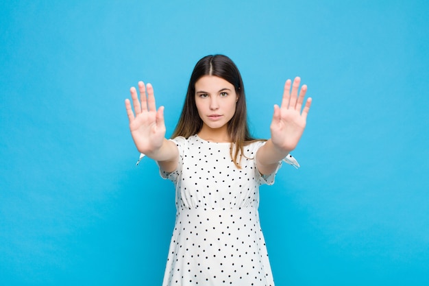 Jovem mulher bonita olhando a entrada proibindo séria, infeliz, irritada e descontente com as duas palmas abertas contra a parede azul