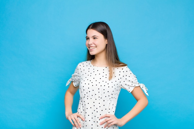 Jovem mulher bonita olhando feliz, alegre e confiante, sorrindo orgulhosamente e olhando para o lado com as duas mãos nos quadris contra a parede azul