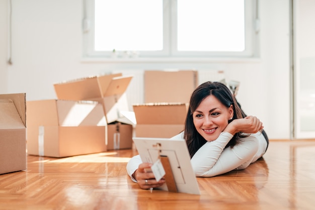 Jovem mulher bonita que encontra-se no assoalho no apartamento novo. Olhando para a moldura pequena.