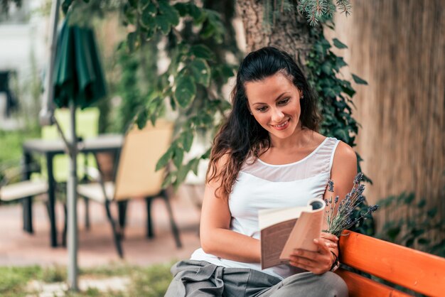 Jovem mulher bonita que lê um livro no banco fora.