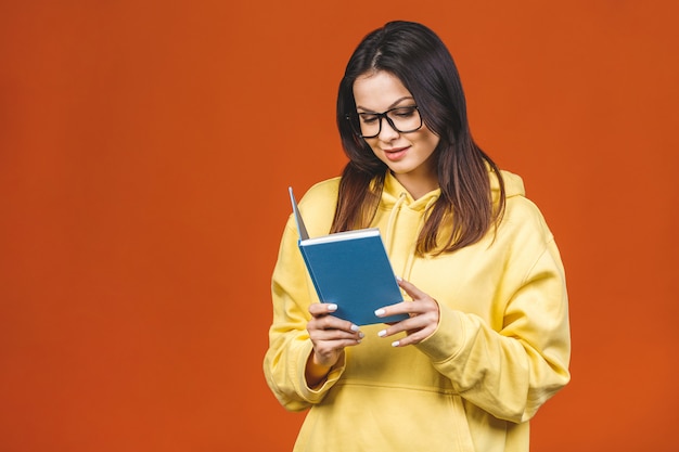 Jovem mulher bonita que veste a posição ocasional isolada sobre o fundo alaranjado, lendo um livro.