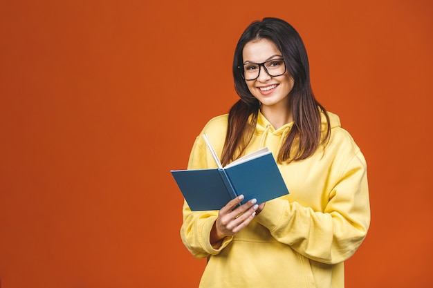 Jovem mulher bonita que veste a posição ocasional isolada sobre o fundo alaranjado, lendo um livro.