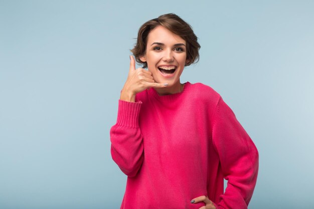 Jovem mulher bonita rindo com cabelo curto escuro no suéter rosa mostrando gesto de telefone enquanto alegremente olhando na câmera sobre fundo azul