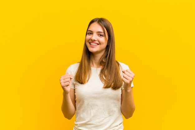 Jovem mulher bonita se sentindo chocado, animado e feliz, rindo e comemorando o sucesso, dizendo uau! contra parede laranja