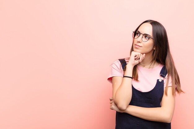 Jovem mulher bonita se sentindo pensativo, pensando ou imaginando idéias, sonhando acordado e olhando para copiar espaço contra parede rosa