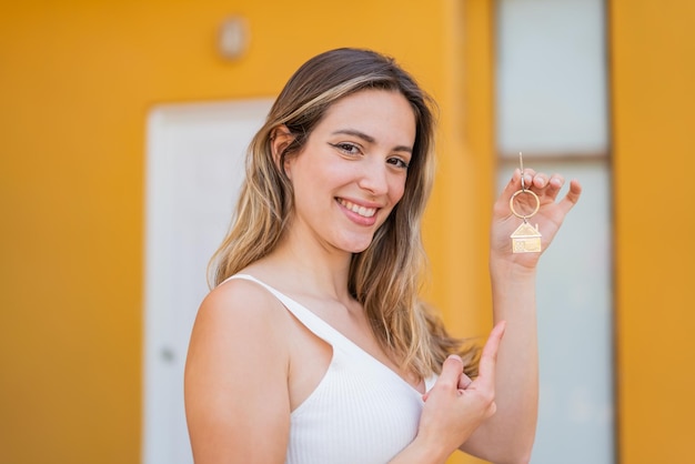 Jovem mulher bonita segurando chaves de casa ao ar livre e apontando para ele