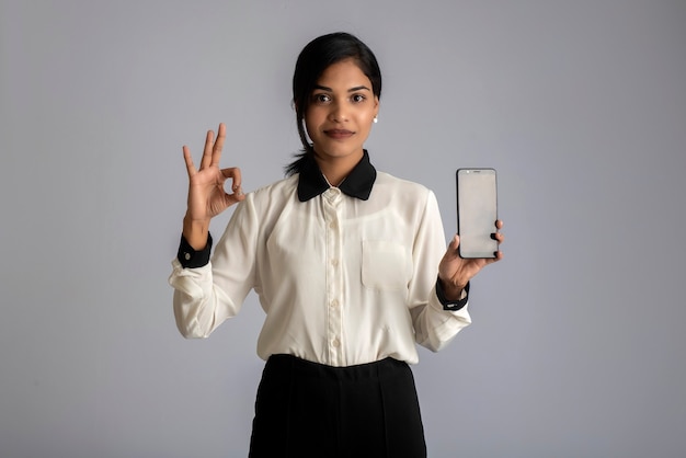 Jovem mulher bonita segurando e mostrando a tela em branco do smartphone ou celular ou tablet em uma parede cinza.