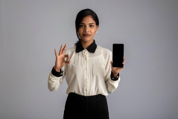 Jovem mulher bonita segurando e mostrando a tela em branco do smartphone ou celular ou tablet em uma parede cinza.