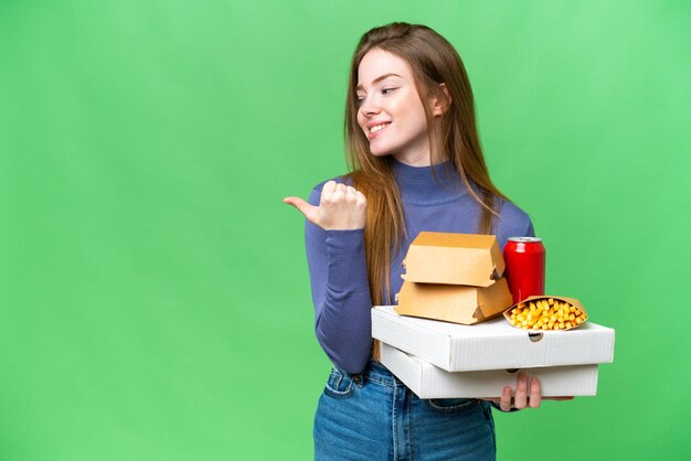 Foto jovem mulher bonita segurando pizzas e hambúrgueres sobre fundo croma chave isolado apontando para o lado para apresentar um produto