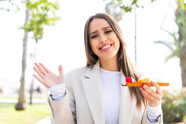 Jovem mulher bonita segurando sashimi ao ar livre saudando com a mão com expressão feliz