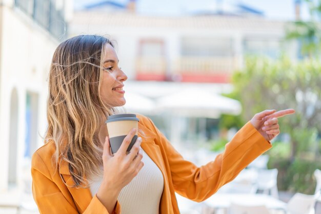 Jovem mulher bonita segurando um café para viagem ao ar livre apontando para o lado para apresentar um produto