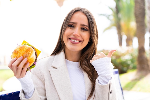 Jovem mulher bonita segurando um hambúrguer ao ar livre apontando para o lado para apresentar um produto