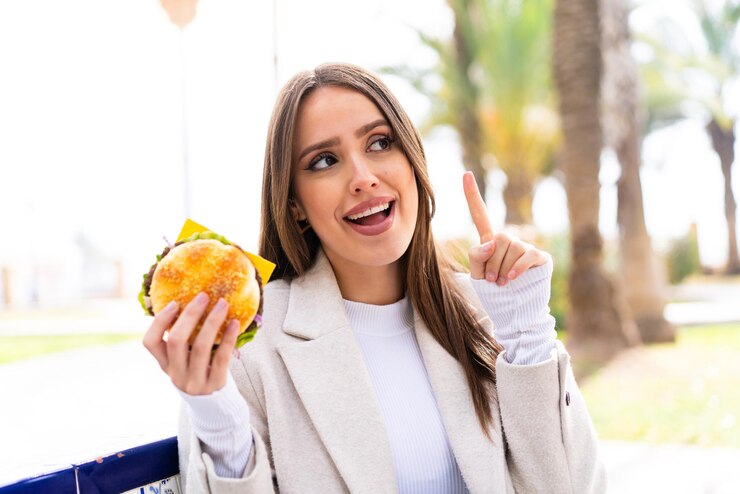 Foto jovem mulher bonita segurando um hambúrguer ao ar livre com a intenção de perceber a solução enquanto levanta um dedo