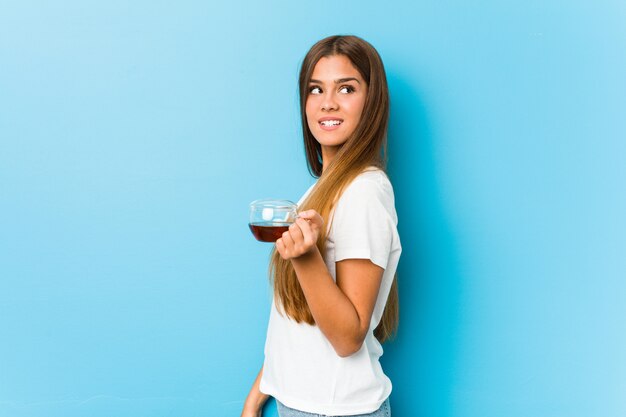 Jovem mulher bonita segurando uma xícara de chá parece de lado sorrindo, alegre e agradável.