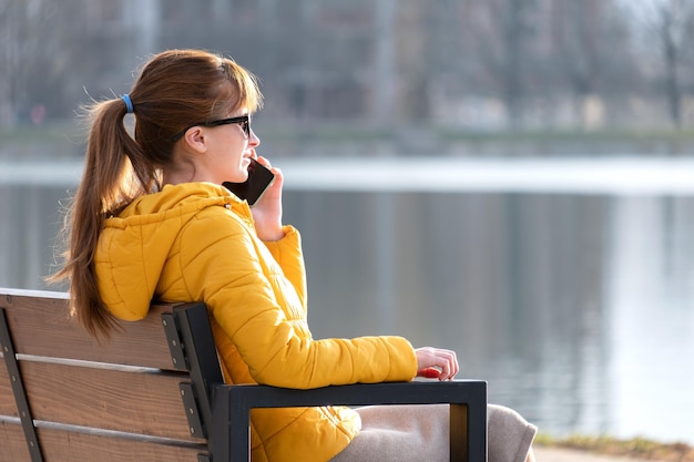 Jovem mulher bonita sentada em um banco de parque falando em seu smartphone ao ar livre na noite quente de outono