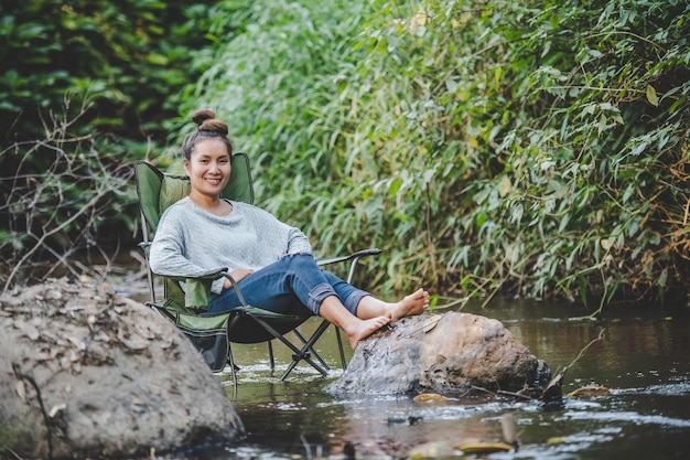 Jovem mulher bonita sentada na cadeira de acampamento no riacho para relaxar ela sorri na floresta natural durante a viagem de acampamento com espaço de cópia de felicidade