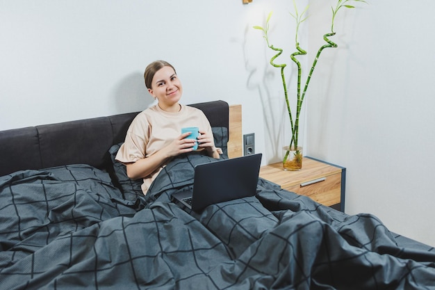 Jovem mulher bonita sentada na cama com café trabalhando com computador em casa trabalho remoto Mulher feliz e sorrindo feliz na cama em casa