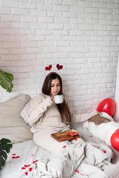 Jovem mulher bonita sentada na cama com seu cachorro comemorando o dia dos namorados comendo doces e bebendo café