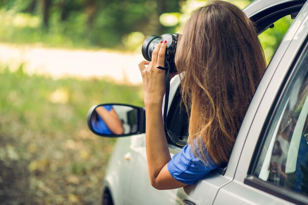 Jovem mulher bonita sentada no carro na floresta e fotografando com câmera digital.