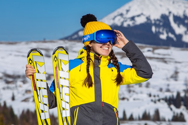 Jovem mulher bonita sorridente segurando esqui. montanhas no fundo. viagens de inverno. copie o espaço