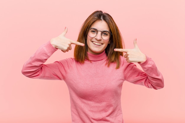 Jovem mulher bonita sorrindo confiante apontando para o próprio sorriso largo, atitude positiva, relaxada e satisfeita contra a parede rosa