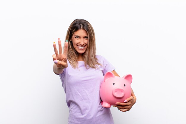 Jovem mulher bonita sorrindo e olhando amigável, mostrando o número dois ou segundo com a mão para a frente, contando com um cofrinho.