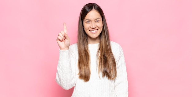 Jovem mulher bonita sorrindo e parecendo amigável, mostrando o número um ou primeiro com a mão em contagem regressiva