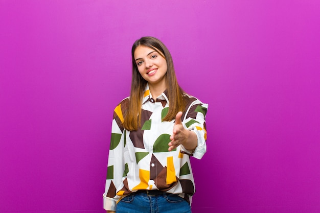 Jovem mulher bonita sorrindo, olhando feliz, confiante e amigável, oferecendo um aperto de mão para fechar um acordo, cooperando sobre parede roxa