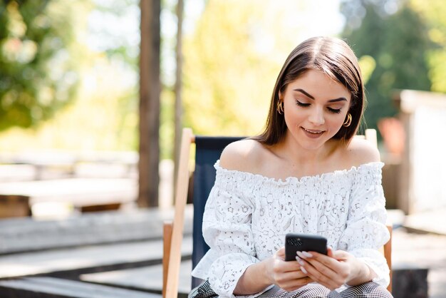 Jovem mulher bonita trabalhando assistindo smartphone na rua do parque moderno, sentada no trabalho remoto, estilo casual de verão