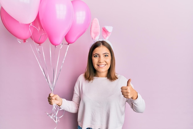 Jovem mulher bonita usando orelhas de coelhinho da páscoa e segurando balões sorrindo feliz e positivo, polegar para cima fazendo excelente e sinal de aprovação