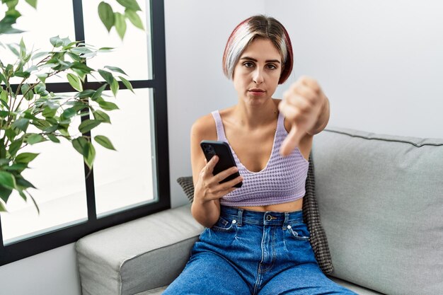 Jovem mulher bonita usando smartphone digitando mensagem sentada no sofá parecendo infeliz e com raiva mostrando rejeição e negativo com expressão ruim de gesto de polegar para baixo
