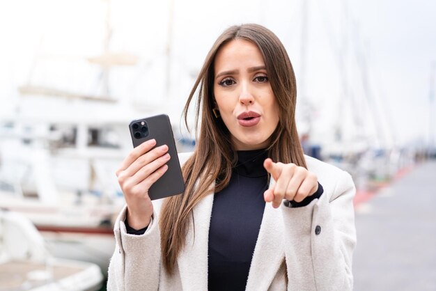 Foto jovem mulher bonita usando telefone celular ao ar livre surpreso e apontando para a frente