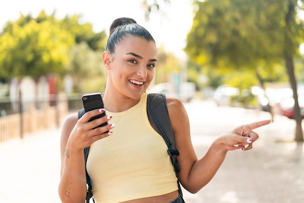 Jovem mulher bonita usando telefone móvel ao ar livre surpreendida e apontando o dedo para o lado