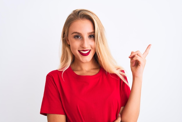 Jovem mulher bonita vestindo camiseta casual vermelha em pé sobre fundo branco isolado com um grande sorriso no rosto, apontando com a mão e o dedo para o lado, olhando para a câmera