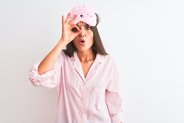 Jovem mulher bonita vestindo máscara de dormir e pijama sobre fundo branco isolado fazendo okey gesto chocado com o olho de rosto surpreso olhando por entre os dedos Expressão incrédula