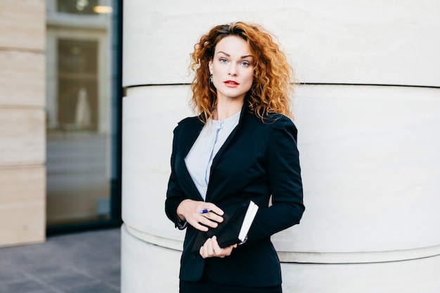 Foto jovem mulher bonita vestindo roupas formais, vestidas formalmente, segurando a bolsa com caneta, tendo um penteado espesso, perto do prédio de escritórios