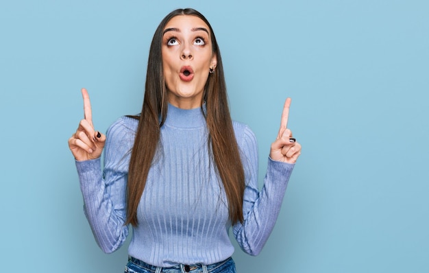 Jovem mulher bonita vestindo suéter de inverno casual espantado e surpreso olhando para cima e apontando com os dedos e braços levantados