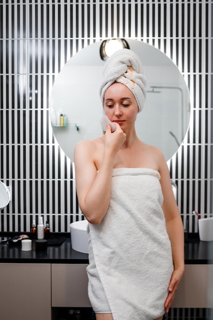 Jovem mulher bonita vestindo toalha branca fazendo massagem facial com raspador de quartzo rosa durante a rotina matinal de beleza no banheiro Tratamentos antienvelhecimento e conceito de cuidados com a pele