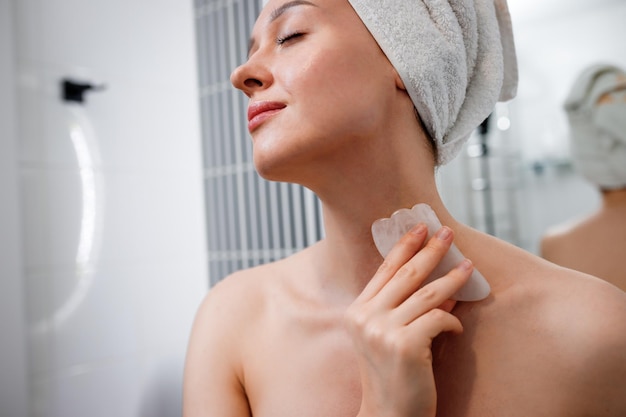 Jovem mulher bonita vestindo toalha branca fazendo massagem facial com raspador de quartzo rosa durante a rotina matinal de beleza no banheiro Tratamentos antienvelhecimento e conceito de cuidados com a pele