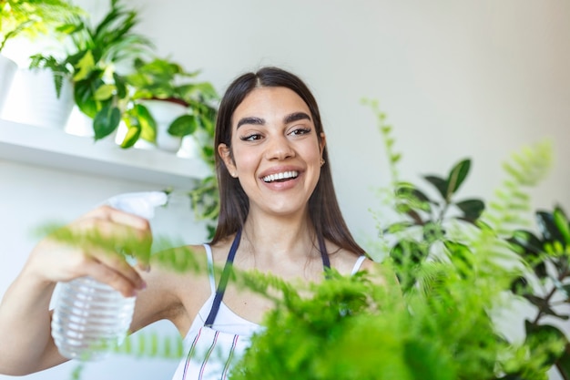 Jovem mulher borrifando uma planta de casa com um borrifador. Jovem alegre aproveita o tempo em casa e rega a planta perto da janela em casa.